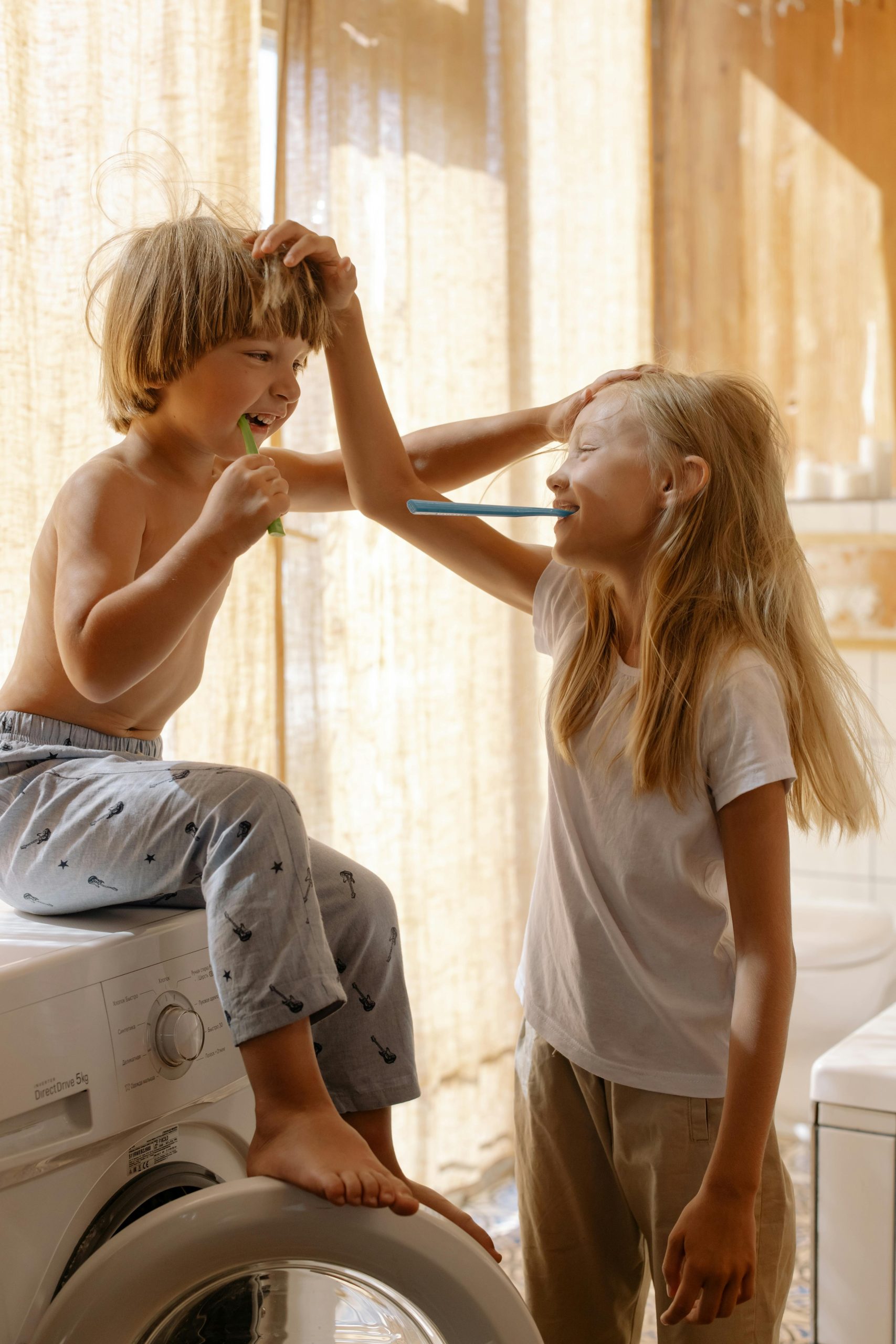 Child sitting on a washing machine brushing teeth with an elder sister’s guidance
